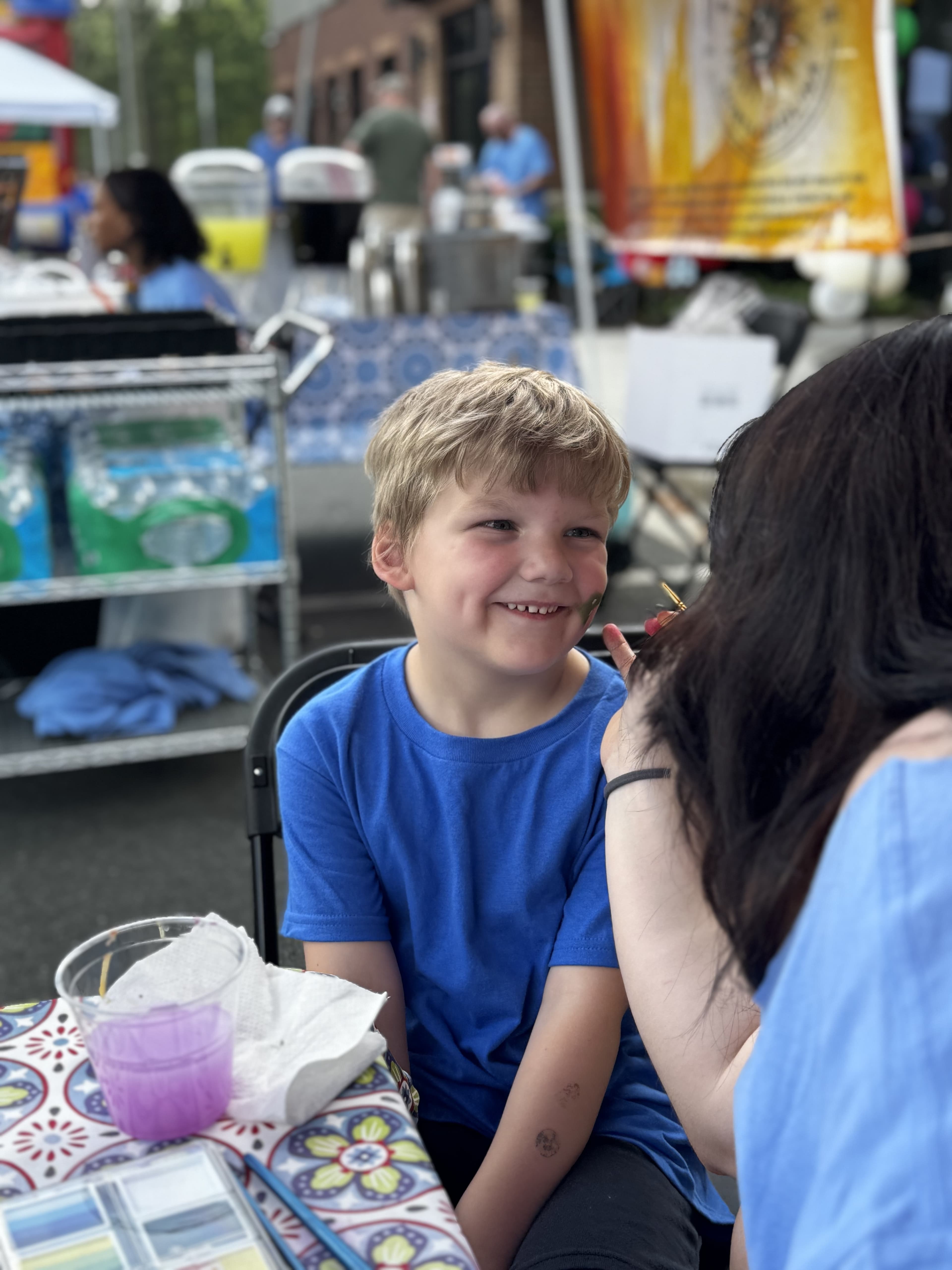 Young boy in a blue shirt smiling while getting his face painted at SmartFest